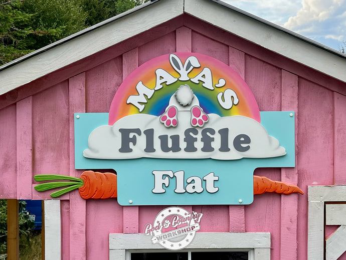 Pink building with a colorful sign reading 'Maya's Fluffle Flat' on a cloudy day at Bark & Bray Sanctuary Farm. Sign by God & Country Workshop in New Hampshire.